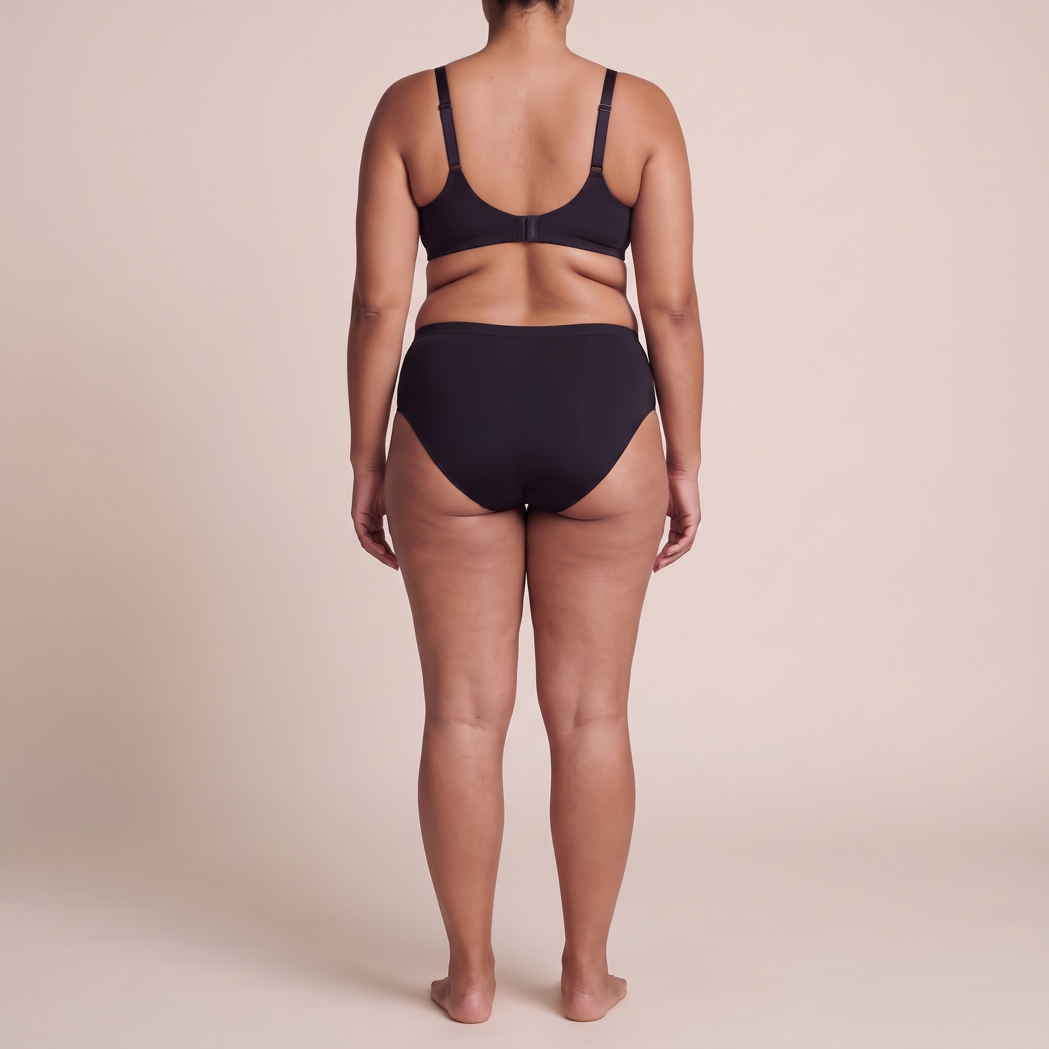 A model shown from the back wearing a matching black two-piece lingerie set, consisting of a bralette with thin shoulder straps and high-cut briefs, against a solid beige studio background.